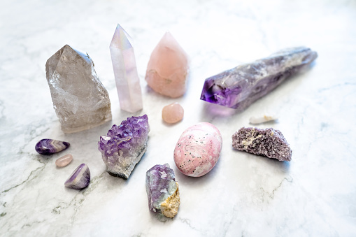 This is a photo of a variety of pink and purple crystals including amethyst, angel aura quartz, rose quartz, smokey quartz, fluorite, lepidolite, and a rhodonite palm stone on a white marble background.