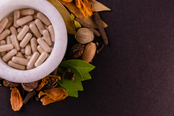 Natural medicine concept, top view of ayurvedic capsules in a mortar and different spices, green leaves on black background with copy space stock photo