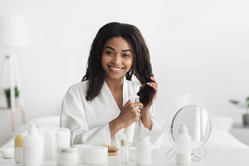 Natural products and cosmetics for hair. Happy african american woman applying essential oil spray on curly hair, sitting in bedroom interior in morning. Split ends repair treatment