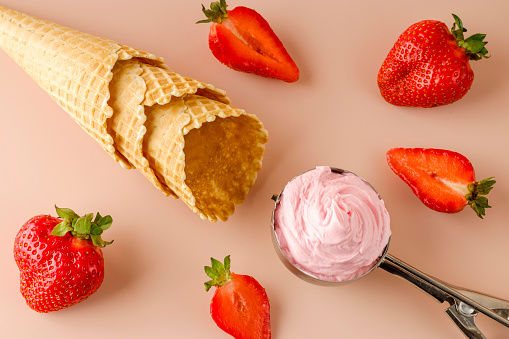 Natural strawberry ice cream in a special spoon. Fresh strawberries and waffle cones on a pink background. Cold summer dessert. Flat lay.