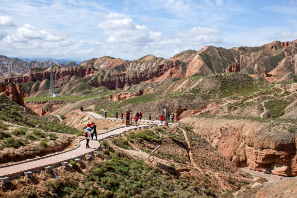Nature Sculptures and Red Sandstone Rocks in the Geopark and Chinese Tourists. Group of Chinese Tourists are Walking Along a Path in Binggou Danxia Canyon Landform in Zhangye, Sunan Region, Gansu Province, China. Red Sandstone Rocks in the Geopark. Clouds and Blue Sky on a Sunny Day. danxia landform stock pictures, royalty-free photos & images