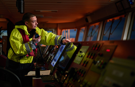 Navigator. pilot, captain as part of ship crew performing daily duties with VHF radio, binoculars on board of modern ship with high quality navigation equipment on the bridge on sunrise.