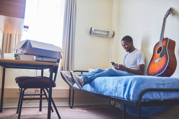 I need to make a call Shot of a cheerful young man texting on his cellphone while being seated on his bed inside of his new home during the day college dorm stock pictures, royalty-free photos & images