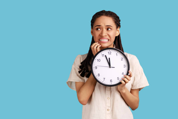 Nervous woman with black dreadlocks biting her nails and holding big wall clock, deadline. stock photo