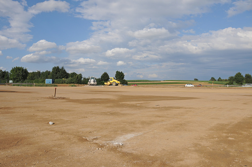 The site is cleared, flattened by bulldozers and ready for development. Building area in the country. Farmland had to yield, the soil will be sealed. The ground has been levelled and prepared for rising a factory or new homes. Wide, empty space. Blue cloudy sky. Trees, bushes, highway and construction machinery in the distance. Horizontal orientation.