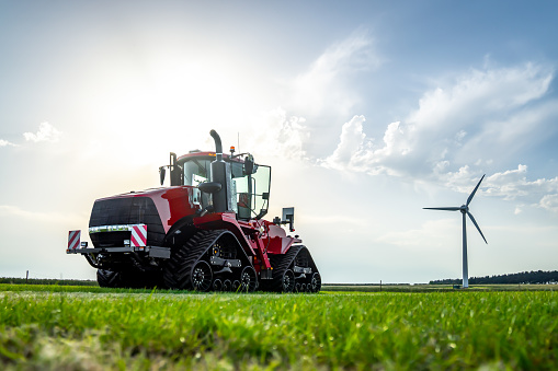 New red modern farm tractor with quad tracks for powerful combine trailer and ploughing trac attachments.  Parked in sunshine on farmers rural agricultural crop field with corn and wind turbine on horizon.  Lush green grass and dramatic low angle shot.