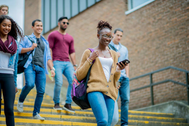 New students on a college campus A woman of African descent walks down the steps of campus with more students walking behind her. She is smiling as she carries a backpack around one shoulder and is holding a phone in her hand. community colleges stock pictures, royalty-free photos & images