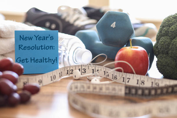 Neujahrsvorsatz, im kommenden Jahr gesund zu werden!  Zu den Objekten gehören: Notiz, Obst, Maßband, Ohrhörer, Sportschuhe, Hanteln, Wasserflasche und Handtuch.   Konzept einer Person, die Gegenstände für eine Übung vorbereitet.