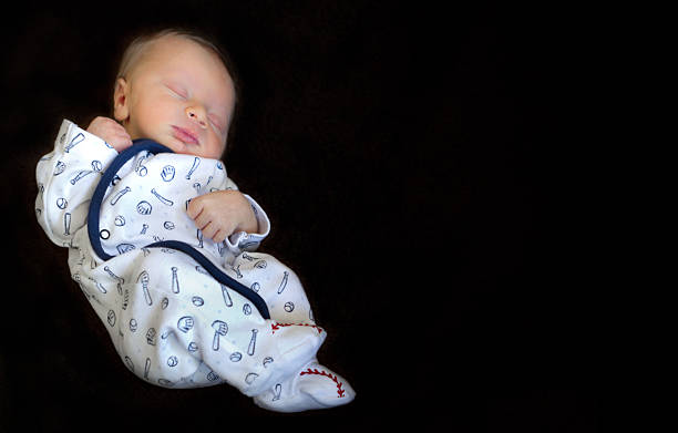 Newborn Baby Isolated on Black Sleeping newborn baby wearing a baseball themed sleeper shown isolated on a black background with plenty of copy space. neicebird stock pictures, royalty-free photos & images