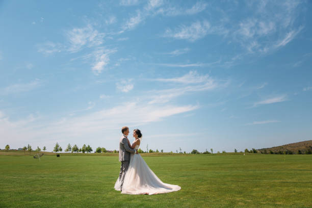 newlyweds are walking along the green field of the golf club on a wedding day. The groom in a business suit is gray and the bride in a luxury white dress with a veil are holding hands stock photo
