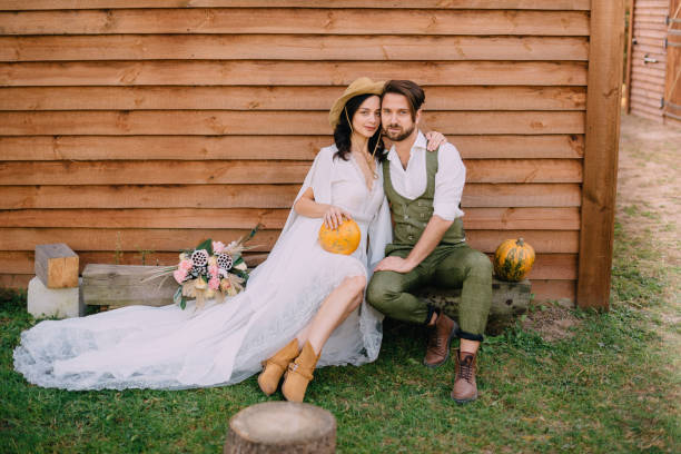 newlyweds dressed in boho style sit near wooden wall stock photo
