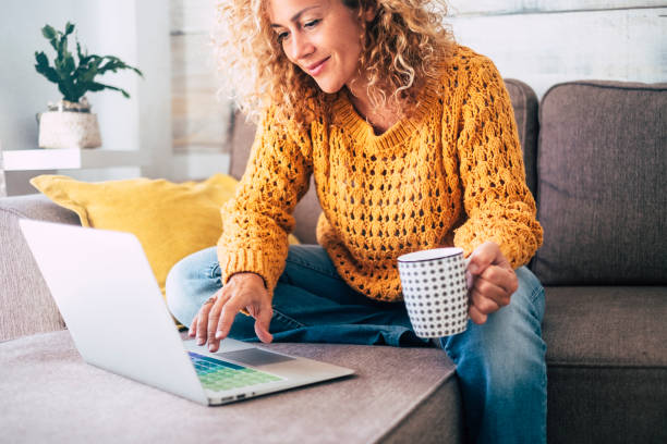 nice beautiful lady with blonde curly hair work at the notebook sit down on the sofa at home - check on oline shops for cyber monday sales - technology woman concept for alternative office freelance - sofá imagens e fotografias de stock
