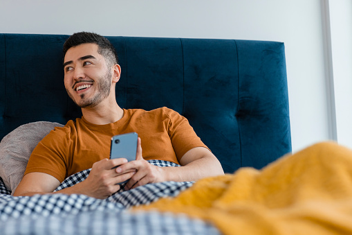 A mid adult Asian man wearing pyjamas in his apartment bedroom on a sunny winters morning. He is lying down and using his smartphone as he relaxes in bed.