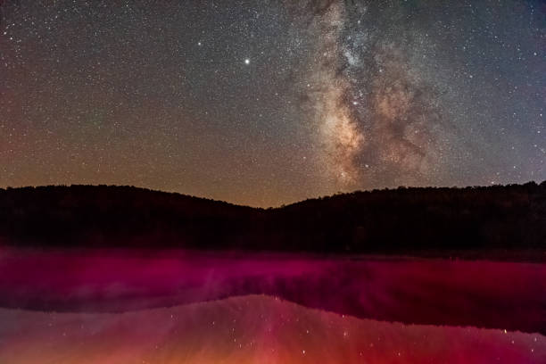 Photo of Night dark sky with milky way galaxy in Spruce Knob Lake in West Virginia with red pink color water reflection from flashlight on fog mist rising steam
