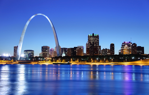The city of St. Louis, Missouri, as seen from across the river.  The Gateway Arch dominates the left upper section of the photo, more than twice as high as any of the buildings in the photo.  In a horizontal line across the background and behind the Gateway Arch are downtown buildings and some skyscrapers.  Lights are just coming on in the buildings even though there is still light in the sky.  A line of beach lies between the buildings and the river, which is a dark blue, mirroring the dark blue of the sky.