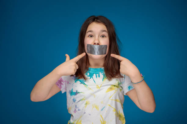 noisy teenager girl silenced with silver tape pointing at her mouth stock photo