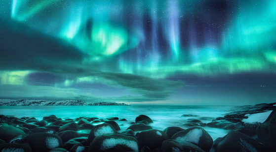 Northern lights. Aurora borealis over ocean in Teriberka, Russia. Starry sky with polar lights and clouds. Night winter landscape with bright aurora, stars, sea, snowy stones in blurred water. Travel