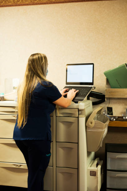 Nurse charting into laptop An blond nurse with long hair wearing navy scrubs and a face mask enters data into her laptop computer on the nursing cart to document patient information during a 12 hour shift inside a medical facility, Midwest, USA inpatient stock pictures, royalty-free photos & images