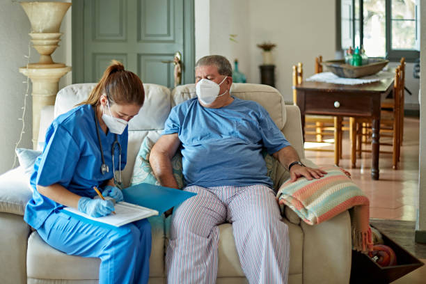 Nurse Making House Call to Care for Senior Man in Recovery Female healthcare worker in early 30s wearing scrubs, protective face mask, and surgical gloves making notes about recovery of senior man during house call. home caregiver stock pictures, royalty-free photos & images