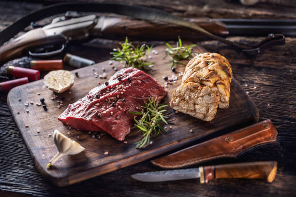 Nutrient rich raw deer venison prepared for a cooking process on a rustic wooden desk with roasted garlic, rosemary and huntig accesories like shot gun and ammunition. stock photo
