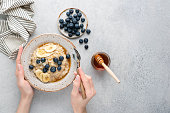 Oatmeal porridge bowl with blueberries, banana, coconut and honey in female hands. Top view copy space for text, grey concrete background. Concept of dieting, clean eating