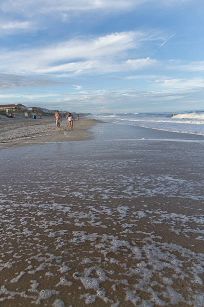 Ocean Tide Converging On Walkers Carolina Beach, Pleasure Island, North Carolina, USA - April 18, 2015: The foamy tide comes in covering the beach in front of people walking along the ocean with blue sky and bright puffy clouds overhead carolina beach north carolina stock pictures, royalty-free photos & images