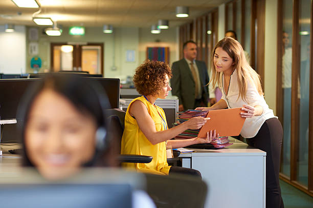 office junior a young office junior takes a file from her supervisor for filing in a large open plan office . Co-workers can be seen defocussed in the foreground and background . secretary stock pictures, royalty-free photos & images