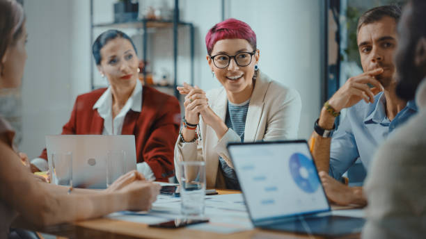 Office Meeting in Conference Room: Beautiful Specialist with Short Pink Hair Talks about Firm Strategy with Diverse Team of Professional Businesspeople. Creative Start-up Team Discusses Big Project stock photo