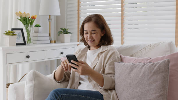 Old adult asia female sit relax on sofa couch smile watch, play, text chat on cellphone at home in happy retired in older people, elderly health care tech life, enjoy pay money online via banking app. stock photo