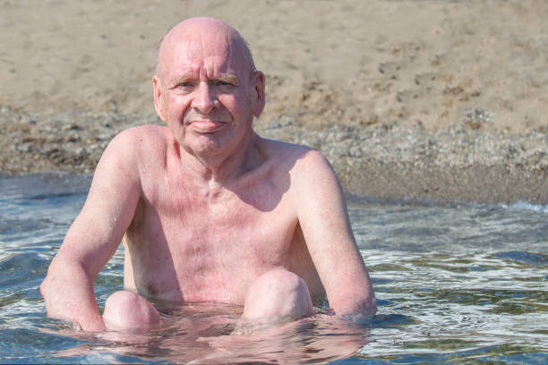 old bald man sits in water sea and sunbathes in sun. sunny autumn day. front view. - bald beach imagens e fotografias de stock