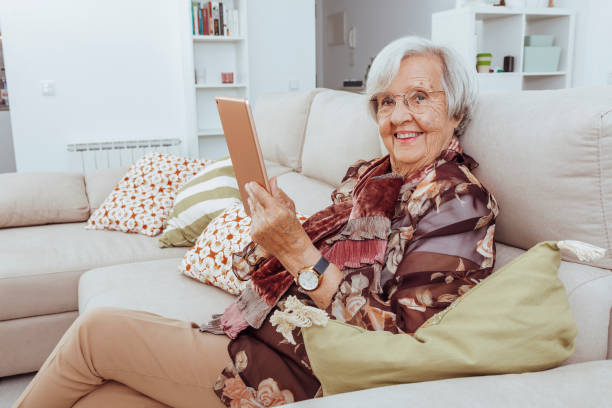 Old grandmother holding digital tablet sitting on couch at home stock photo