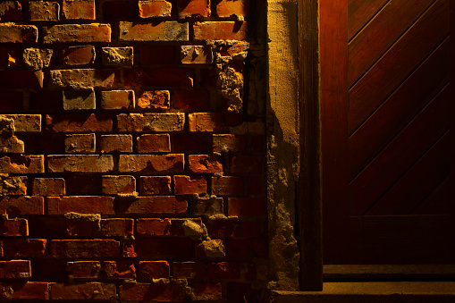 Old grungy and damaged red brick wall at night beside a wooden door with light of a lantern shining on it