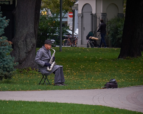 New York, USA - July, 2017: old man in a raincoat and a hat playing saxophone in a park.
