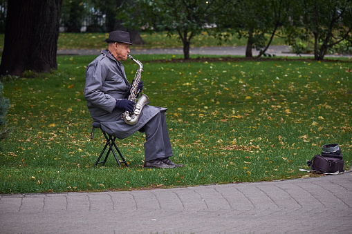 New York, USA - July, 2017: old man in a raincoat and a hat playing saxophone in a park.