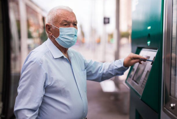 Old man in mask using cash machine on tram station stock photo