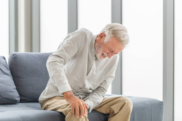 Old man suffering from knee pain sitting sofa in the living room, Elderly man suffering from knee pain while sitting on the sofa stock photo