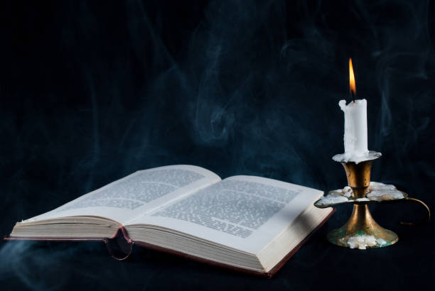 Old open book and candle in candlestick and smoke on dark black background stock photo