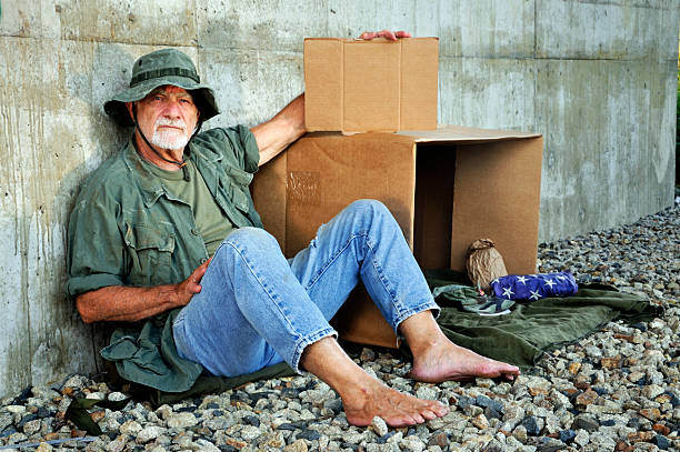 Old Soldier Looks at Camera Holding Blank Sign A Vietnam war veteran (actual) appears to be down and out. In this shot he bears a serious expression while holding a blank cardboard sign for statements like "Will Work for Benefits," etc. Scene staged. mike cherim stock pictures, royalty-free photos & images