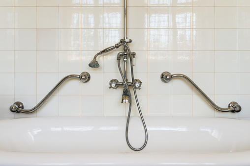 Old stylish retro bathtub with a traditional shower faucet in a white tiled bathroom - with metal handles for the elderly