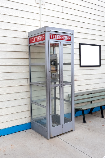 Vertical shot of an old telephone booth with a blank sign on the wall next to it on the right side.  There is a bench below the sign.