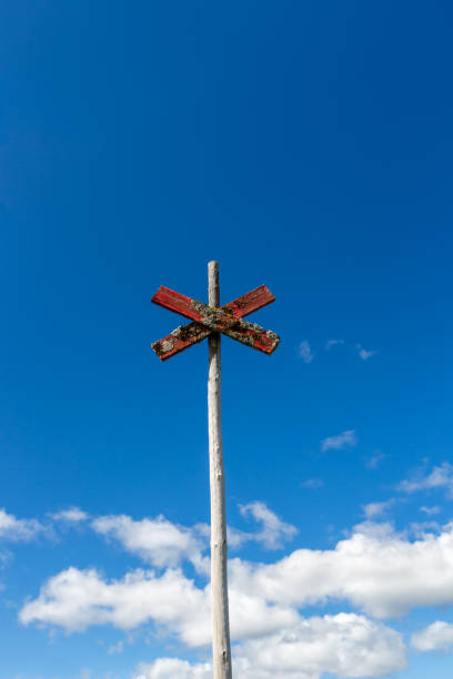 Old weathered wooden mountain trail sign post with red cross against blue sky with clouds. stock photo