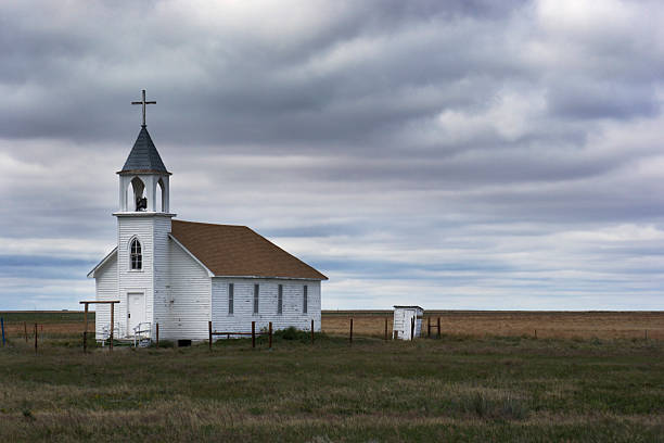 Old White Wooden Church in Rural Field Scene with Storm Horizontal view of a lonely rural church by a prairie farm field, under a stormy sky in western rural South Dakota, U.S.A. The old white wooden built structure has a steeple with a cross and bell tower. The forbidding landscape, cloudscape and Christian symbols reflect dark simplicity and loneliness. bell tower tower stock pictures, royalty-free photos & images