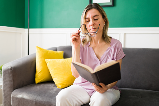 Attractive mature woman with eye sight problems using her glasses to read a book at home