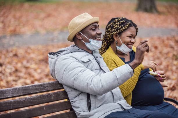 op een mooie herfstdag, zittend op een bankje in het park, een paar neemt een pauze van het dragen van hun beschermende maskers. - pregnant couple outside stockfoto's en -beelden