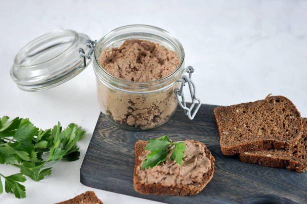 On a wooden board, several slices of black bread with liver pate. Close-up. On a wooden board, several slices of black bread with liver pate. Next to a glass jar with pate. In the frame, green parsley. Light background.  Close-up. Macro photography. liver pâté photos stock pictures, royalty-free photos & images