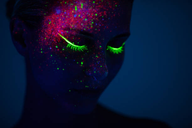One woman painted with fluorescent make up One young woman painted with fluorescent colors standing in front of ultraviolet light. Space is dark with blue background. cosmetics stock pictures, royalty-free photos & images