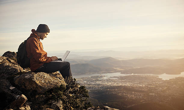 Online in the open air Shot of a young hiker using his laptop while sitting on top of a mountain extreme terrain stock pictures, royalty-free photos & images