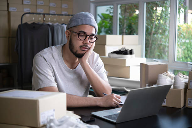 Online seller entrepreneur in a warehouse looking at laptop and worrying about e-commerce business due to lack of sales and customers. stock photo