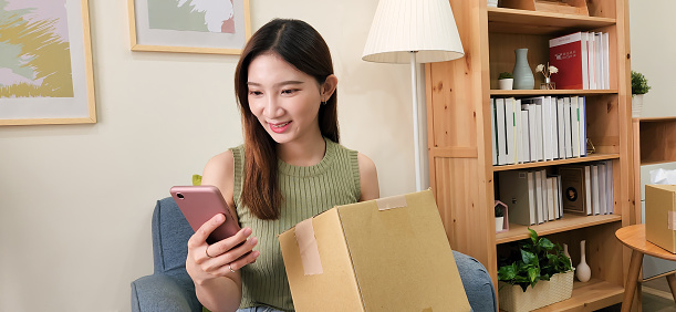 asian woman look package use phone confirm information on parcel from online shopping delivery service sit sofa at home