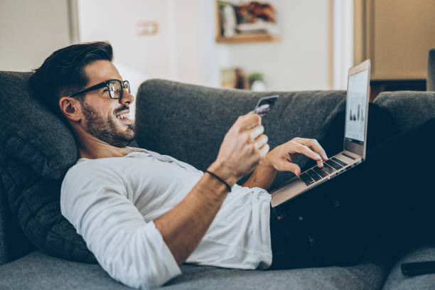 Online shopping Smiling man lying on the couch and shopping online with credit card and laptop online shopping stock pictures, royalty-free photos & images